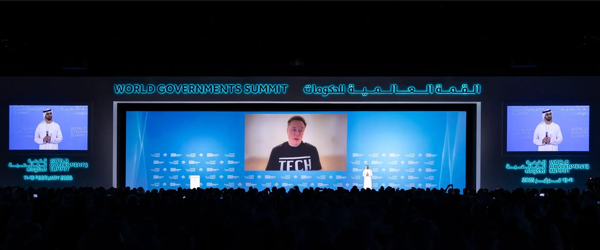 إيلون ماسك يلقي كلمة عبر اتصال مرئي في القمة العالمية للحكومات بدبي، يظهر على الشاشة الكبيرة بقميص مكتوب عليه TECH، بحضور مسؤولين إماراتيين وجمهور غفير.
